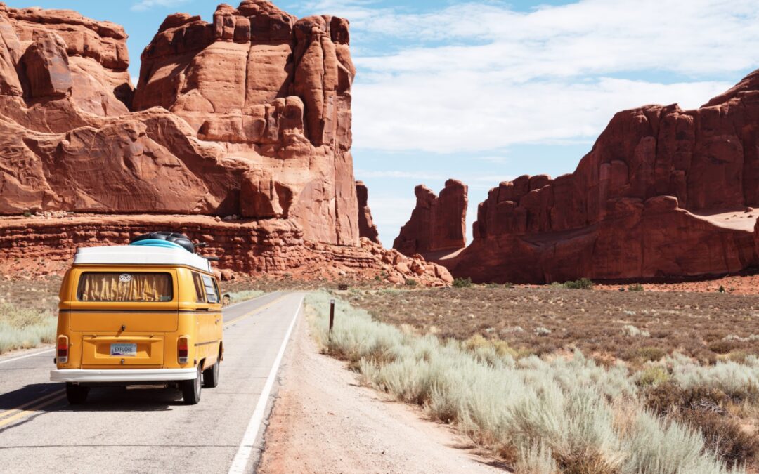 Retro yellow Volkswagen style camper van driving down a desert highway with large red rocks ahead